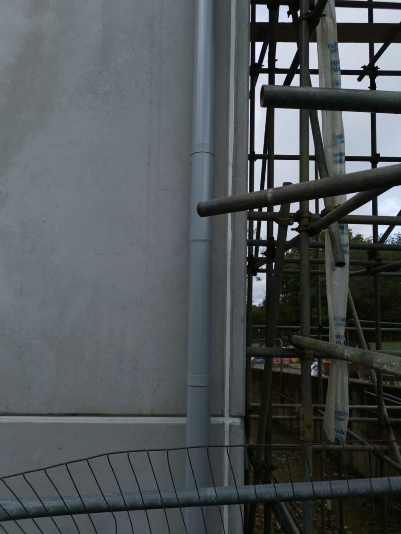 Construction scaffolding beside a gray concrete wall, with a vertical drainage pipe attached to the wall.