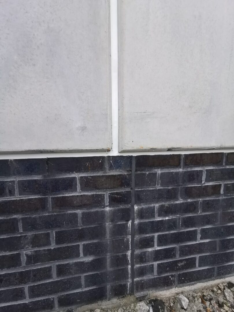 Exterior wall with light gray concrete panels above a dark brick foundation; a vertical seam runs between panels in the middle.