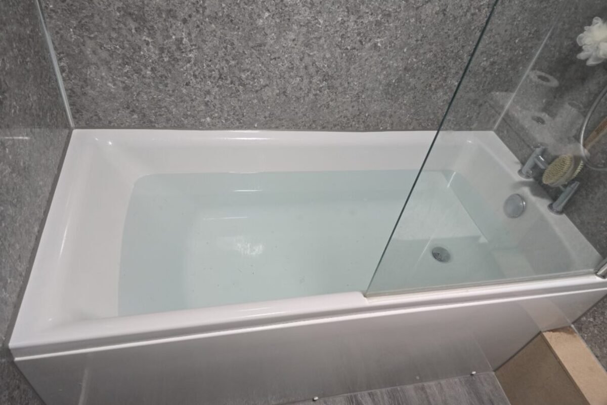 White bathtub with a clear glass shower screen in a gray stone-tiled bathroom wall setting.
