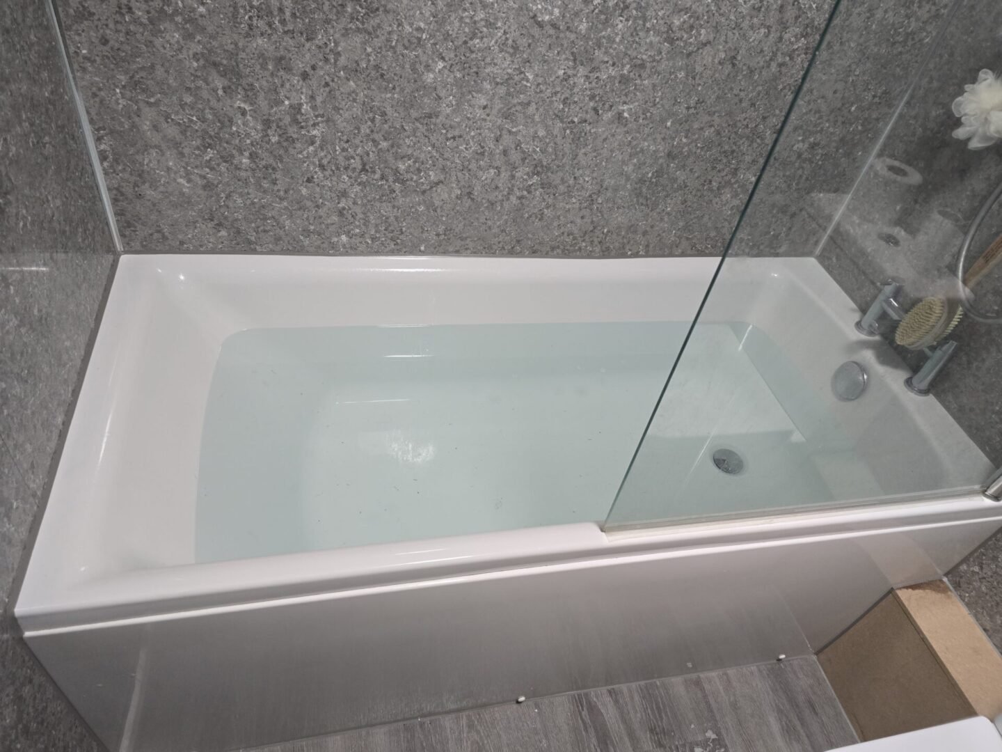 White bathtub with a clear glass shower screen in a gray stone-tiled bathroom wall setting.