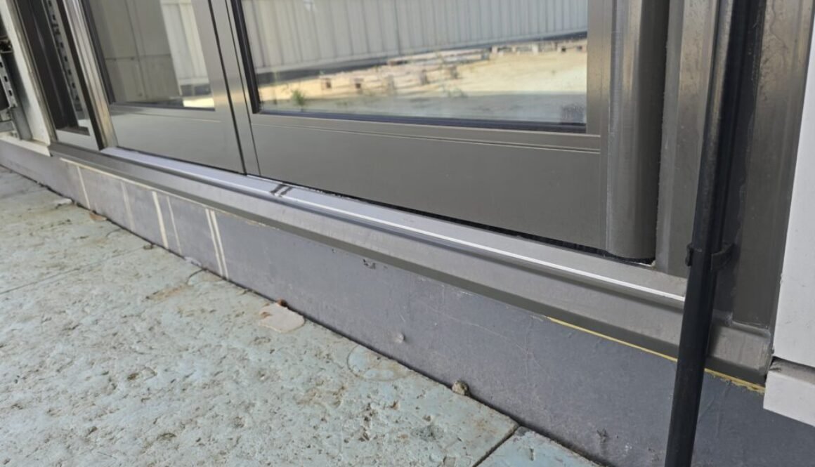 Close-up of a dark gray metal-framed sliding glass door at ground level, showing the bottom sill and a concrete exterior surface nearby.