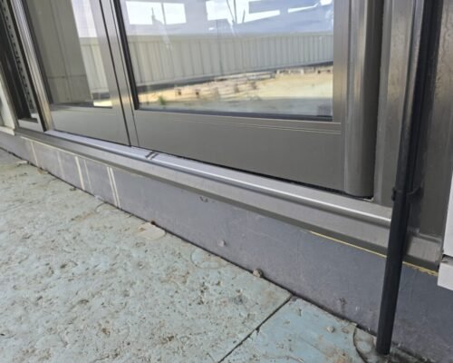 Close-up of a dark gray metal-framed sliding glass door at ground level, showing the bottom sill and a concrete exterior surface nearby.
