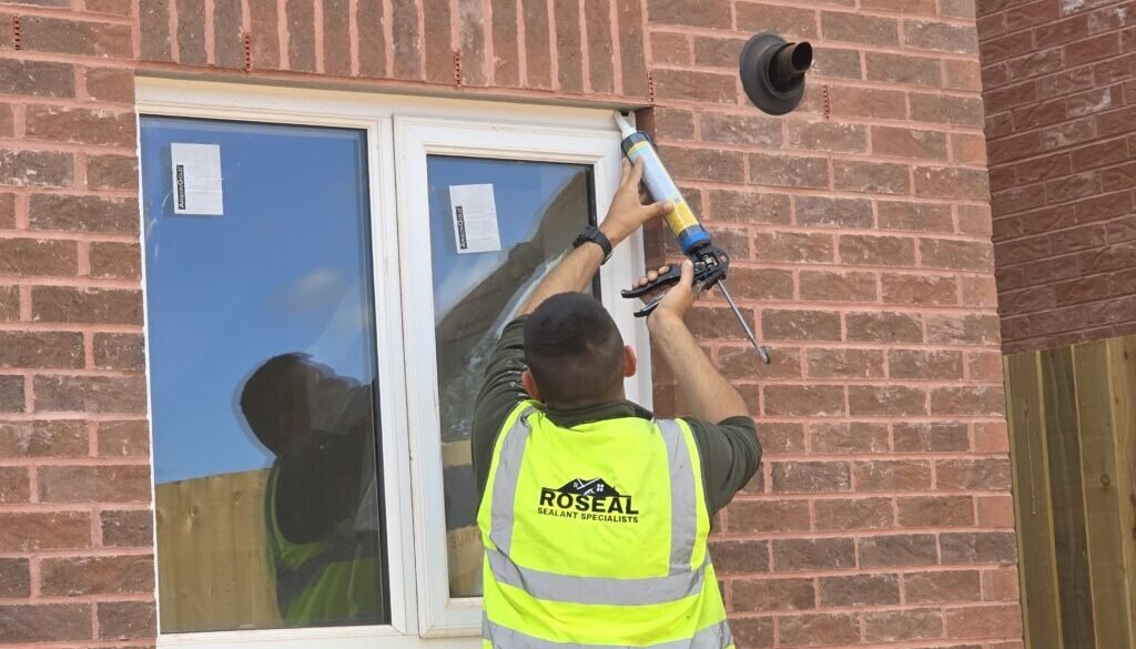 Worker in a neon safety vest seals the edge of a window on a brick house using a caulking gun.