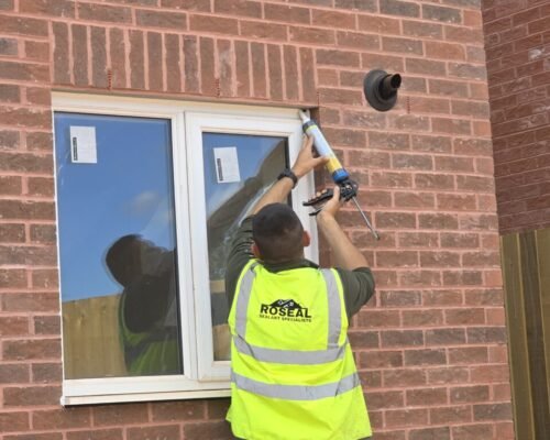 Worker in a neon safety vest seals the edge of a window on a brick house using a caulking gun.