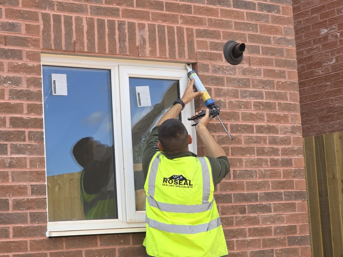 Worker in a neon safety vest seals the edge of a window on a brick house using a caulking gun.